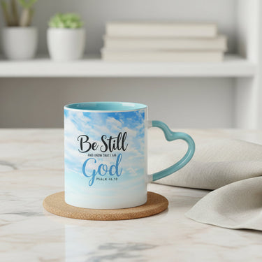 A ceramic mug with a sky-blue and white watercolor design, featuring the text 'Be Still in the Presence of God Psalm 46:10' and a heart-shaped handle, styled on a white wooden table with a small potted succulent and a folded napkin.