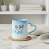 A ceramic mug with a sky-blue and white watercolor design, featuring the text 'Be Still in the Presence of God Psalm 46:10' and a heart-shaped handle, styled on a white wooden table with a small potted succulent and a folded napkin.
