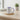 A white ceramic mug with a purple handle and interior, featuring the printed text 'I WILL NEVER Leave YOU FOR FORSAKE YOU JOSEPH 1:9' and purple floral illustrations. The mug sits on a round cork coaster on a light wood table, with a blurred background showing a window, books, and a potted plant.