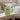 A light green ceramic mug with a bright green handle sits on a light wood bedside table in a softly lit bedroom. The mug has a printed message that reads, 'I'M A CHILD OF GOD' with 'God' in a green circle.