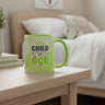 A light green ceramic mug with a bright green handle sits on a light wood bedside table in a softly lit bedroom. The mug has a printed message that reads, 'I'M A CHILD OF GOD' with 'God' in a green circle.
